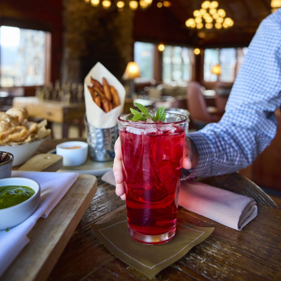 A person holding a glass of red cocktail with mint leaves on top, seated at a wooden table in what appears to be a restaurant.