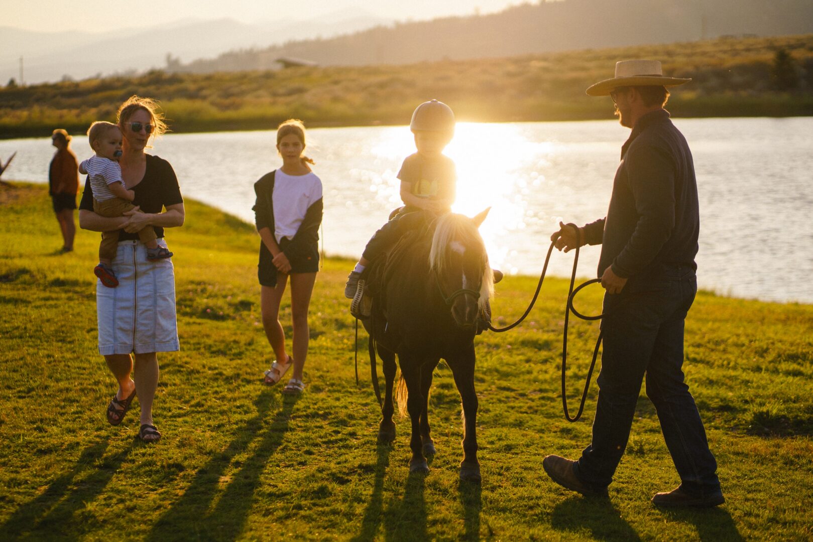 Family playing outside with one kid riding a pony as the sunsets with a pond in the background.