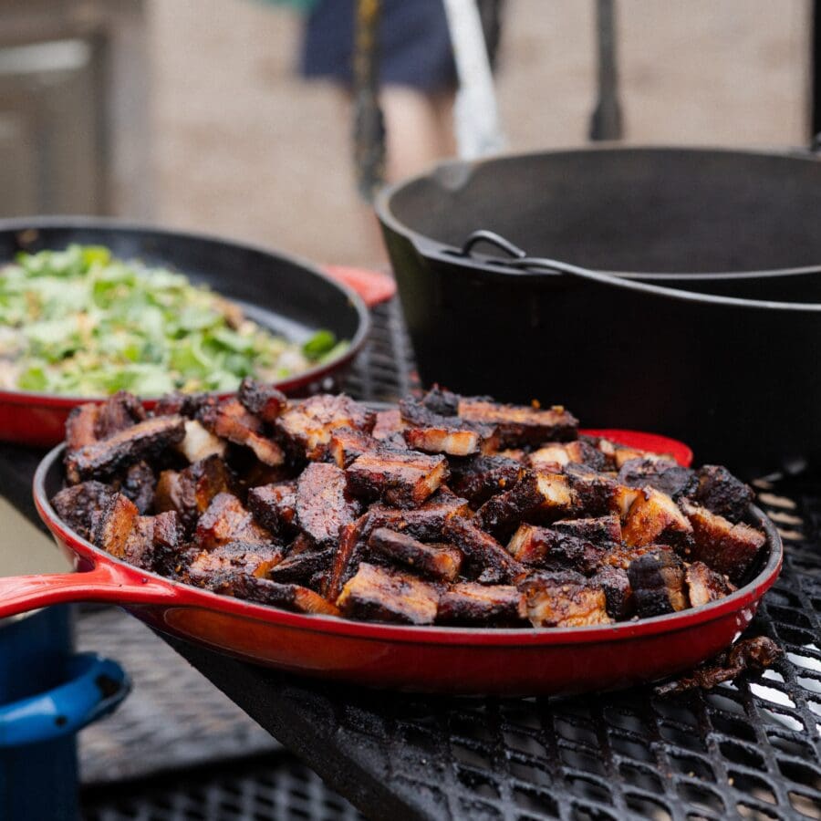 Different meats and vegetables being cooked in a cast iron skillet over a fire.