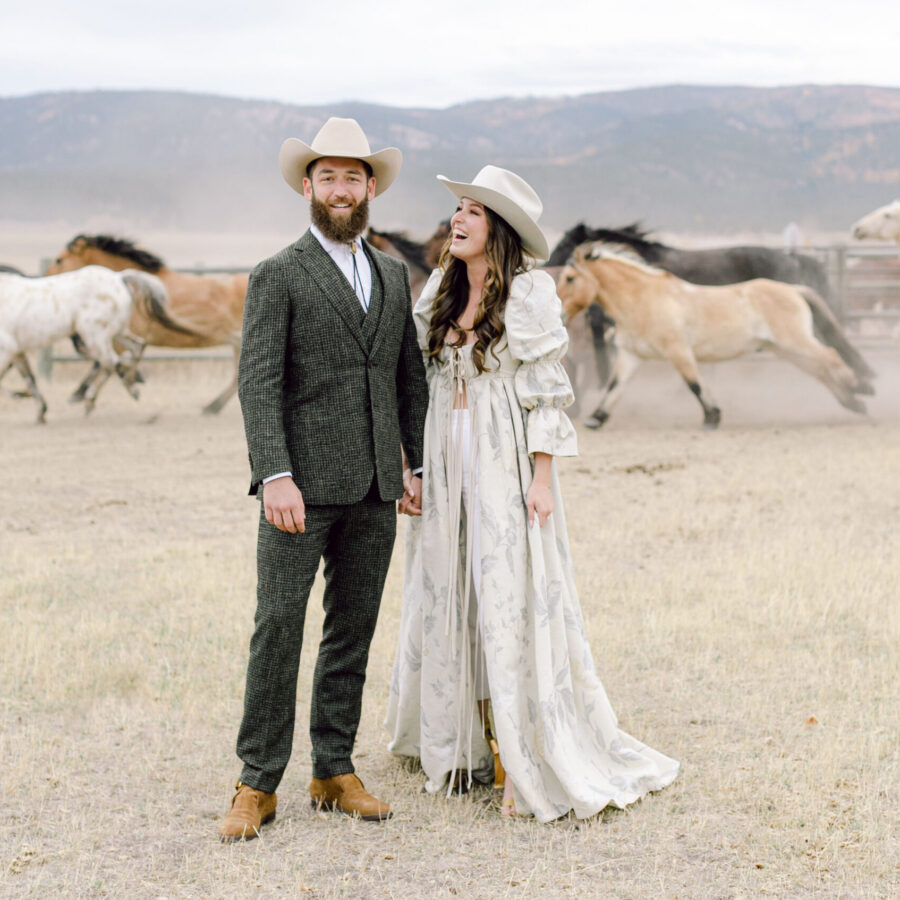 A bride and groom in western style wedding clothes standing outside with horses running behind them.